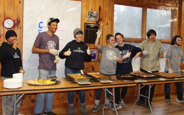 A group of people stand behind a long table lined with food, appearing to be preparing to serve during the family seminar of an outward bound intercept course.
